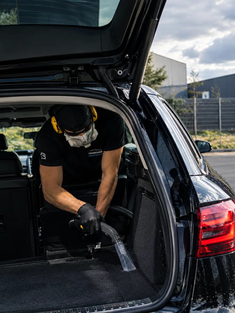 Technik v chráničích sluchátek vysává koberec v kufru černého kombi